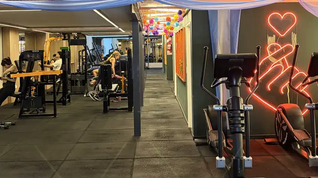 Sala de musculación con máquinas a la izquierda, un pasillo con pompones de colores al fondo y elípticas junto al neón del corazón.